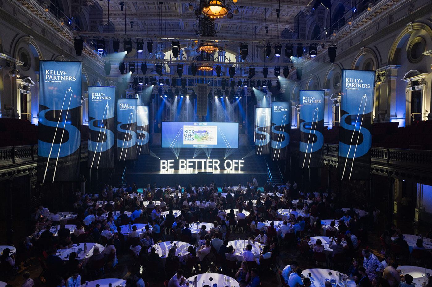 Kelly+Partners Kick Off 2025: a full hall of tables facing a stage lit with Be Better Off.